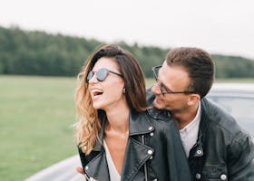 Young couple laughing together outdoors in stylish jackets near a black car, embracing with joy.